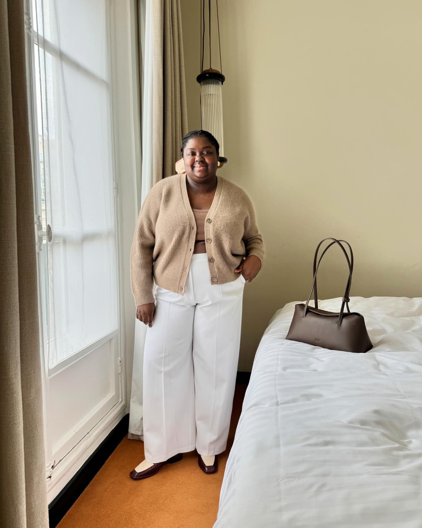 British plus size influencer Abisola Omole poses in a modern bedroom wearing a neutral outfit with a tan cardigan, camel top, white pants, brown minimalistic bag, and two-tone loafers