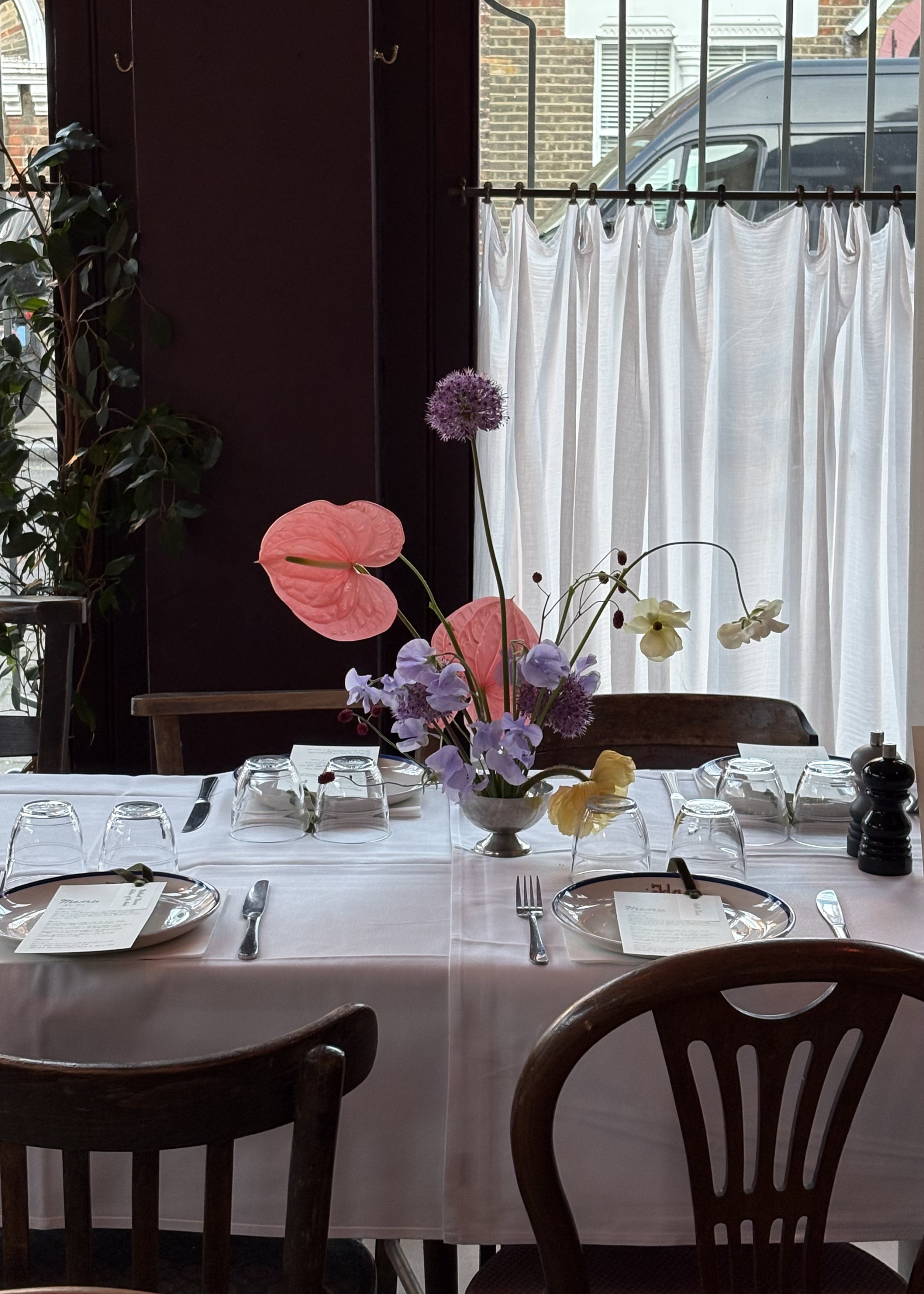A restaurant table with mismatched wood chairs, a modern ikebana arrangement, and place settings