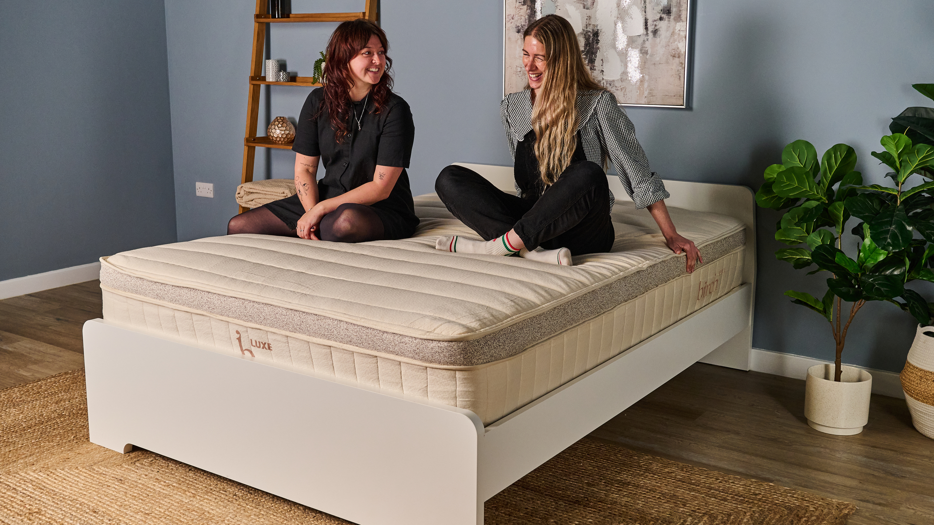 Two Tom&#039;s Guide mattress testers sat crossed legged on the Birch Luxe Natural mattress laughing and smiling