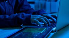 Cyber crime concept image showing hacker typing on keyboard in dimly-lit room with tablet pictured on desk.