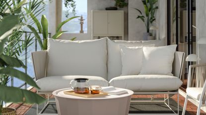 A two seat conversation set with a round coffee table on a balcony on top of a dark gray rug surrounded by foliage, with dark-framed sliding doors leading inside next to a garden closet