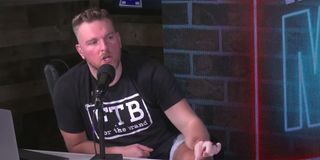 Pat McAfee sitting at a desk with a black shirt on and talking into a microphone.