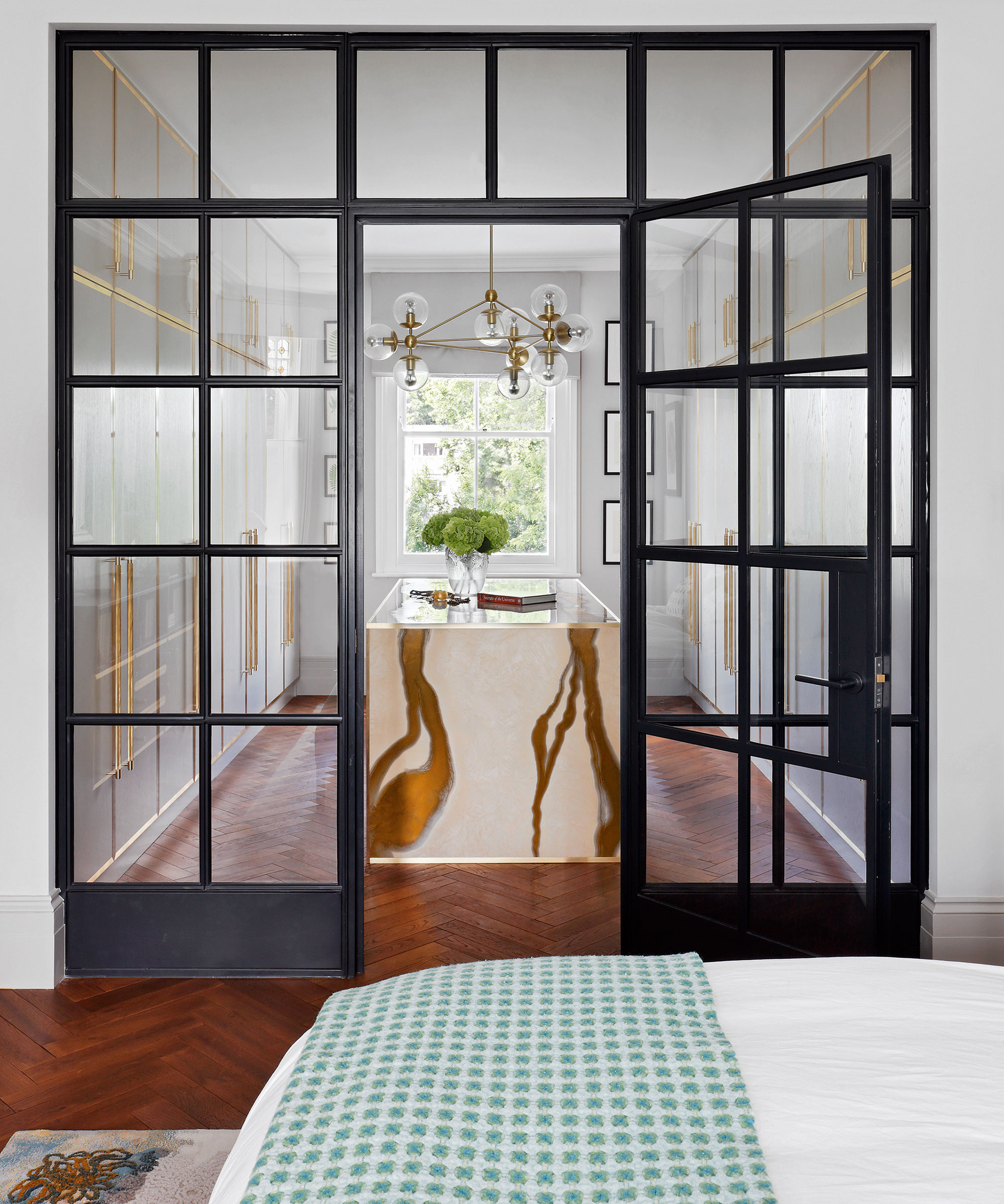 Walk-in closet ideas with a walk-in wardrobe separated from the main bedroom by glass crittall-style doors, with wooden flooring and a chandelier.