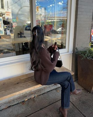 Content creator Mobina Peiman poses wearing a brown sweater, dark wash jeans, and brown boots.