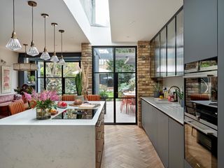 kitchen extension with brickwork and crittall glazing. In the space is a kitchen island with a stove top and plants. Above the island are light pendants.