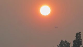 Photo of the sun shrouded in a reddish brown haze. The outline of a helicopter can be seen flying through the hazy sky.
