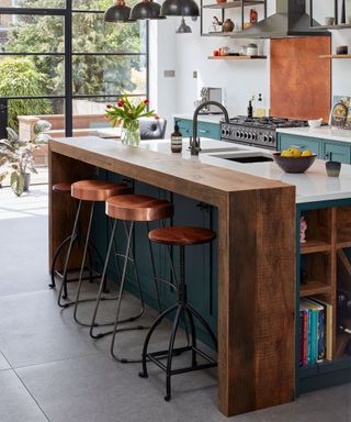 Modern kitchen with kitchen island with wooden and marble countertops, metallic bar stools with copper seats, blue painted island base and cabinets, bookshelf integrated into island, black hanging pendant lights