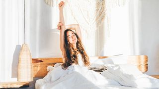 A woman with long brown wavy hair stretches and smiles upon waking up early in the morning and feel energized and ready for the day ahead