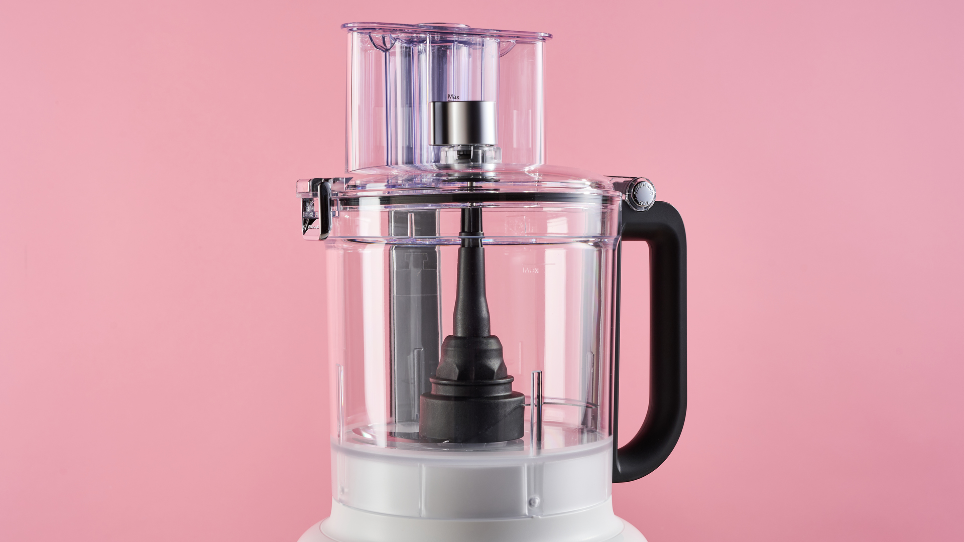 A close up of the 13 cup work bowl of the KitchenAid 13 cup food processor, the black handle is on the right hand side. It is photographed against a pink background.