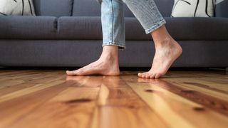 Cropped shot of female legs walk barefoot on wooden warm floor near couch in living room at home