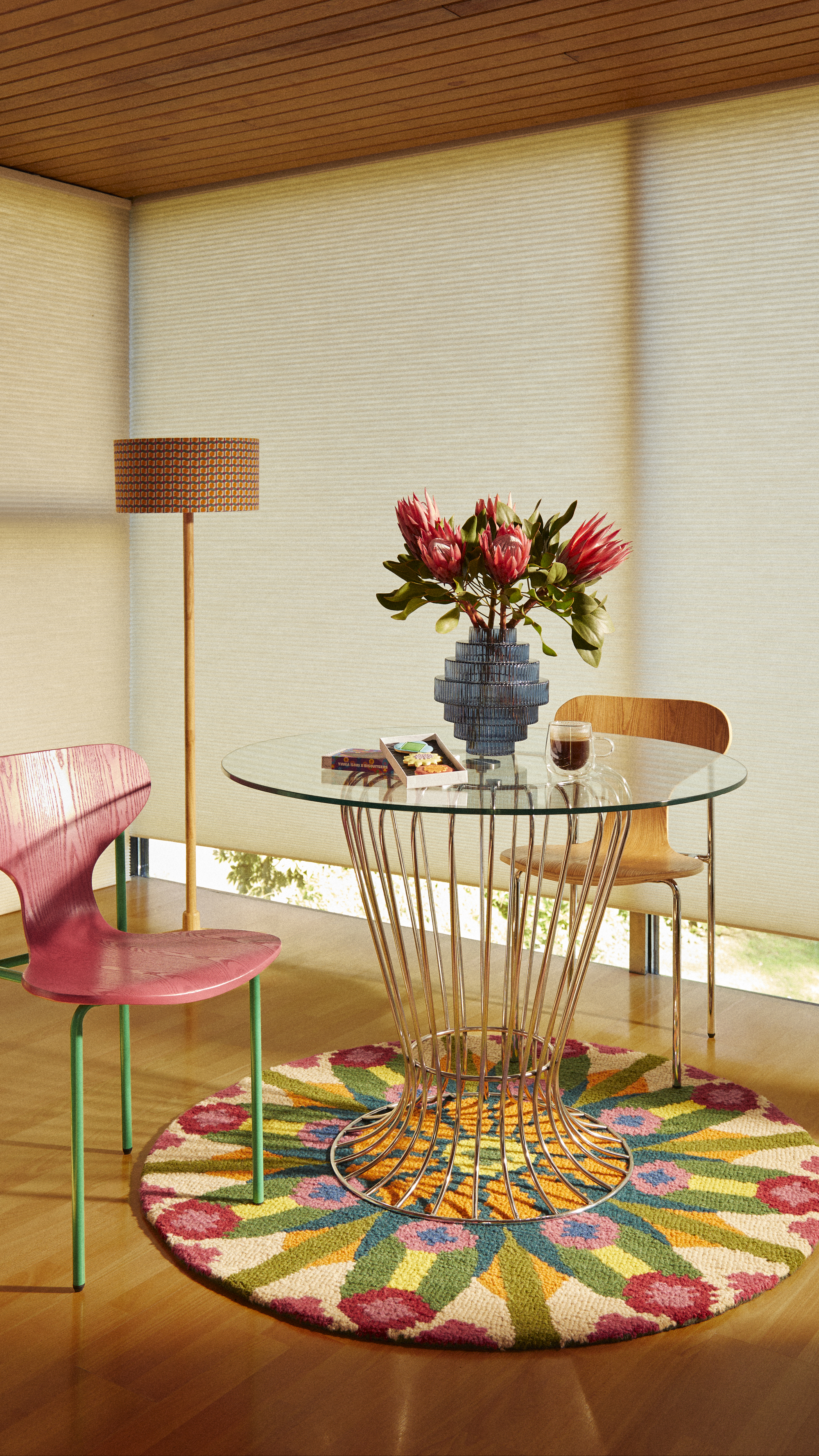 round glass table with steel legs, sheer blinds, timber ceiling, floor lamp, colourful round rug under the table, and a pink and green chair