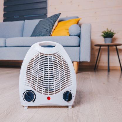 A small electric fan heater on the floor in a living room in front of a light blue sofa with dark blue and yellow cushions