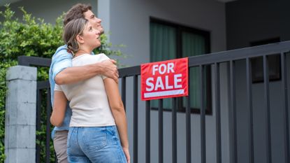 A young couple stands with their arms around each other looking at a house with a For Sale sign