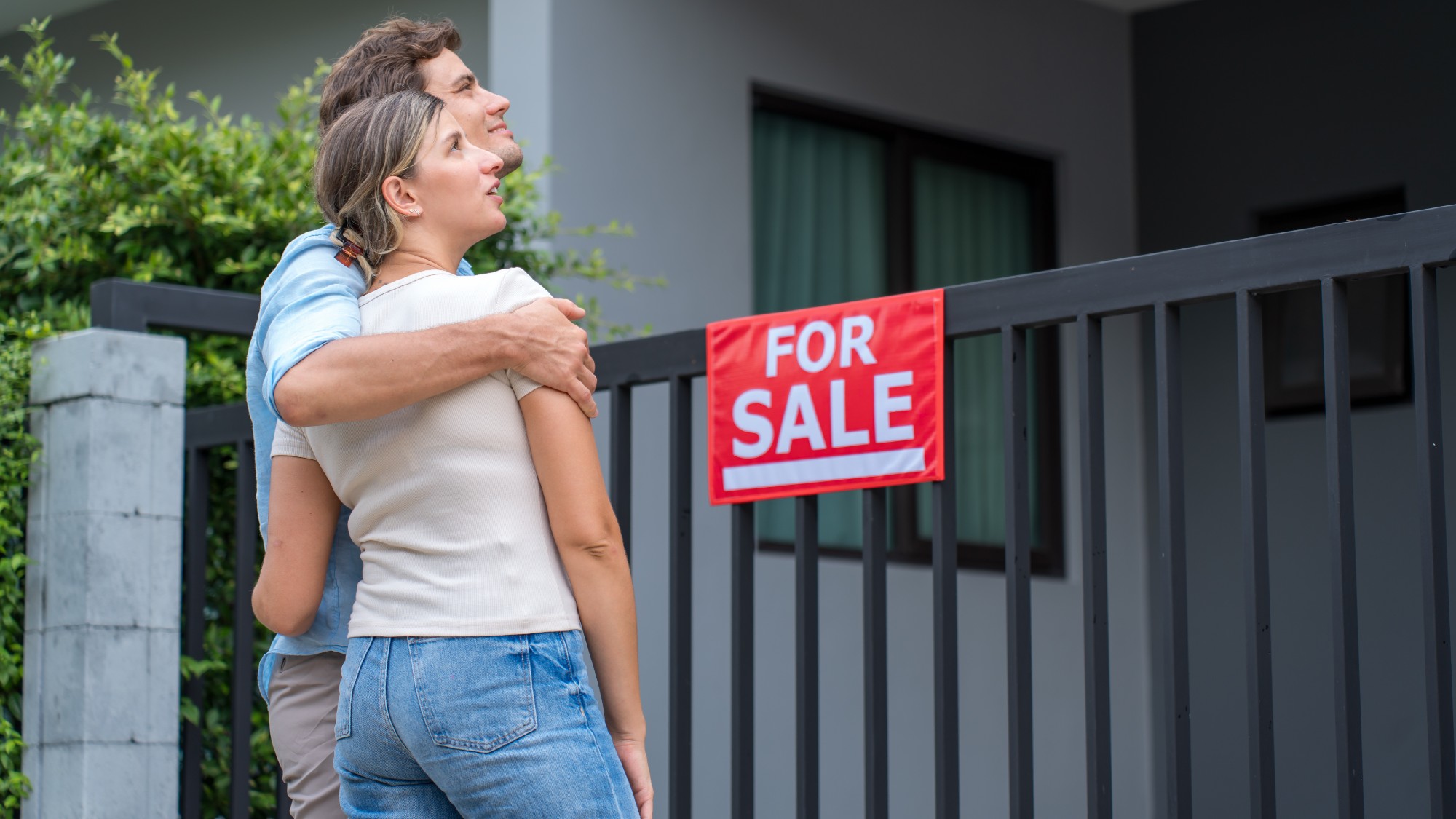 A young couple stands with their arms around each other looking at a house with a For Sale sign