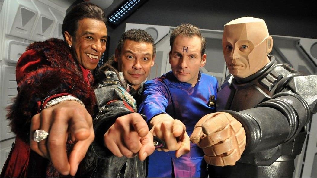 Still from the British sci-fi sitcom called Red Dwarf. Here we see four of the main cast pointing at the camera. From left to right: The Cat, Lister, Rimmer, and Kryten.