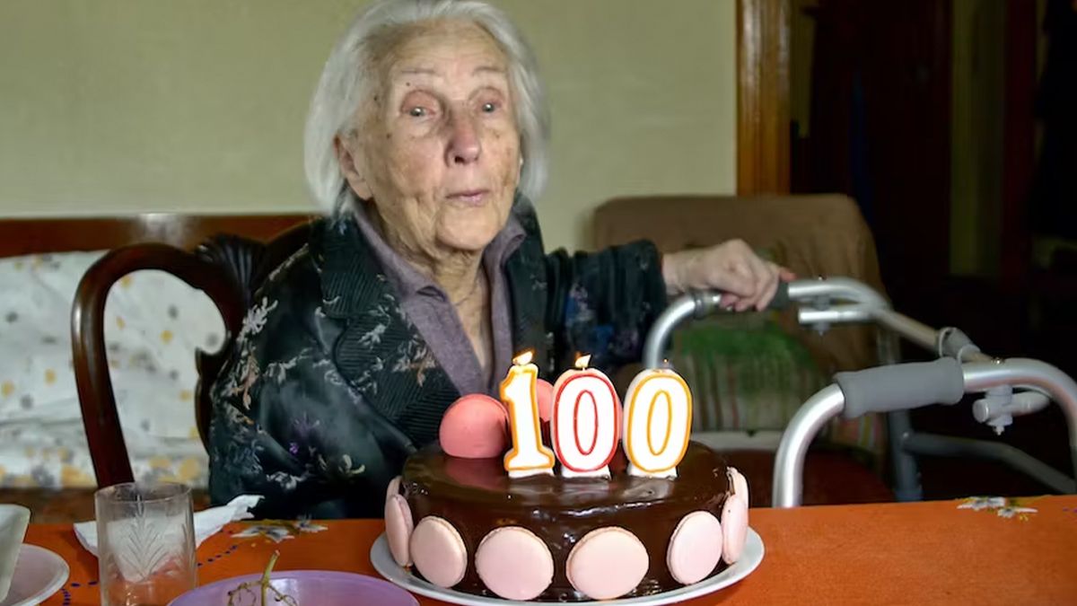 Older woman sitting at a table prepares to blow out candles shaped like the number &quot;100&quot; on top of a chocolate and pink cake.