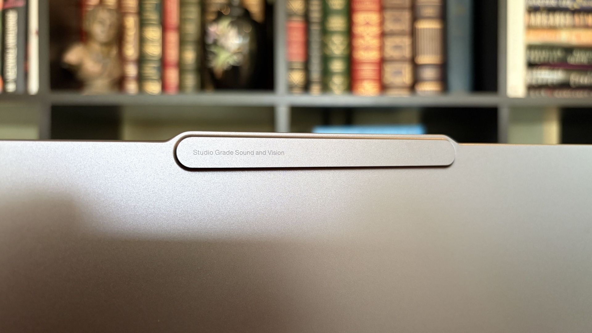 A close-up of a Lenovo Pro 9n laptop lid with a narrow, pill-shaped logo plate, set against softly blurred bookshelves; the scene feels quiet and refined.