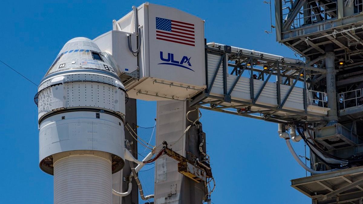 a rocket with a cone-shaped spacecraft on top. it is beside a launch tower. a crew access arm reaches from the tower to the spacecraft with the word &quot;ULA&quot; and an american flag on the side