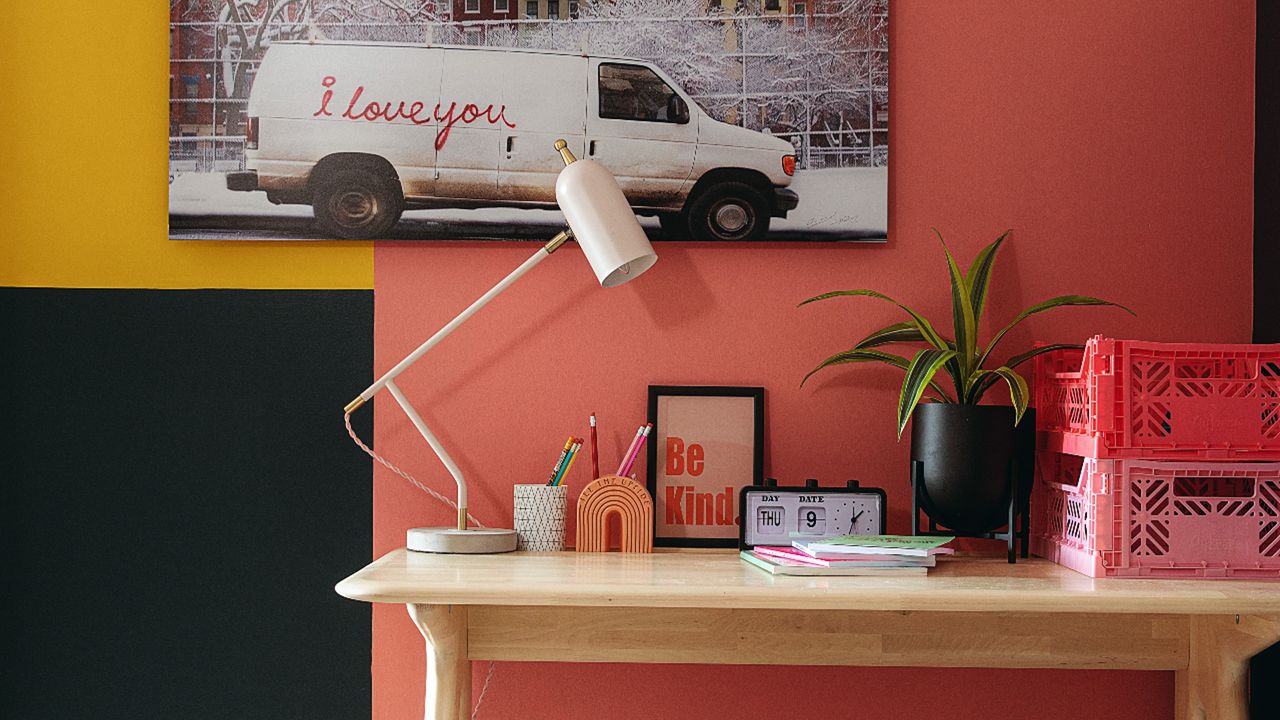 Pink, yellow, and green painted wall, wall art, wooden desk with houseplants, crates, lamp, and decorative items