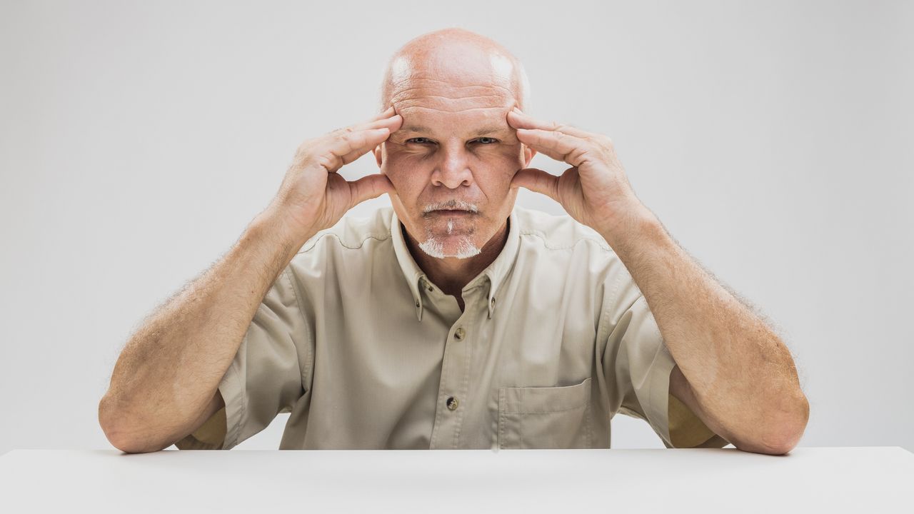 A man sitting at a table presses his fingertips to his temples like he&#039;s a mind reader.