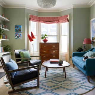 mid century style living room with green walls, blue sofa, black and wood armchairs, coffee table, blue and cream graphic rug, antique side board, retro red floor lamp, red patterned blinds, bay window, open shelving,