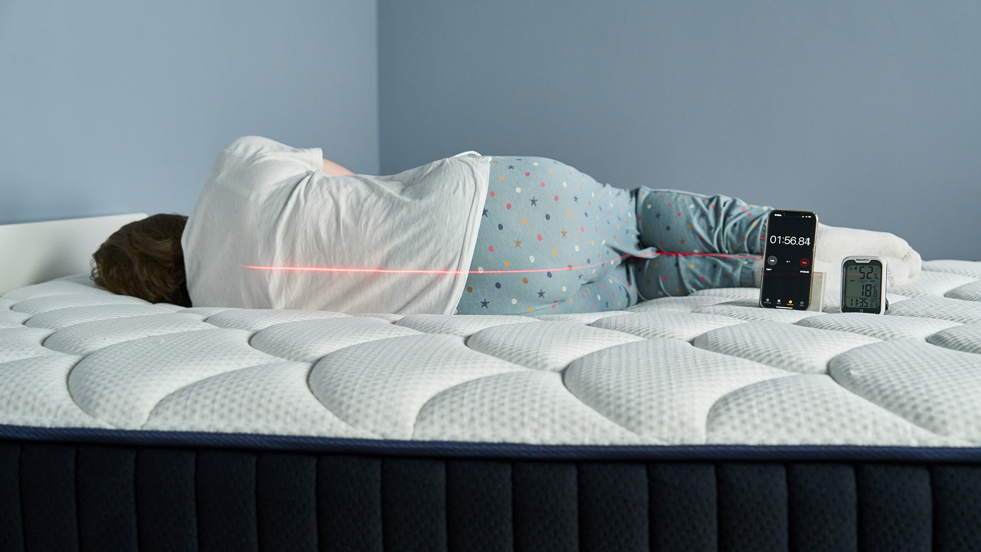 This image shows our mattress tester Ruth Jones lying on her side on the DreamCloud Classic Hybrid Mattress with her back to the camera in our Sleep Lab. A laser line indicates optimal spine position. Next to her is a phone showing a stopwatch and a thermostat