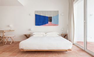 Interior view of the room in the Casa Mãe hotel. Large, airy space, with floor-to-ceiling windows, a large bed with white linen, and modern painting above it in blue, gray, and pink tones.