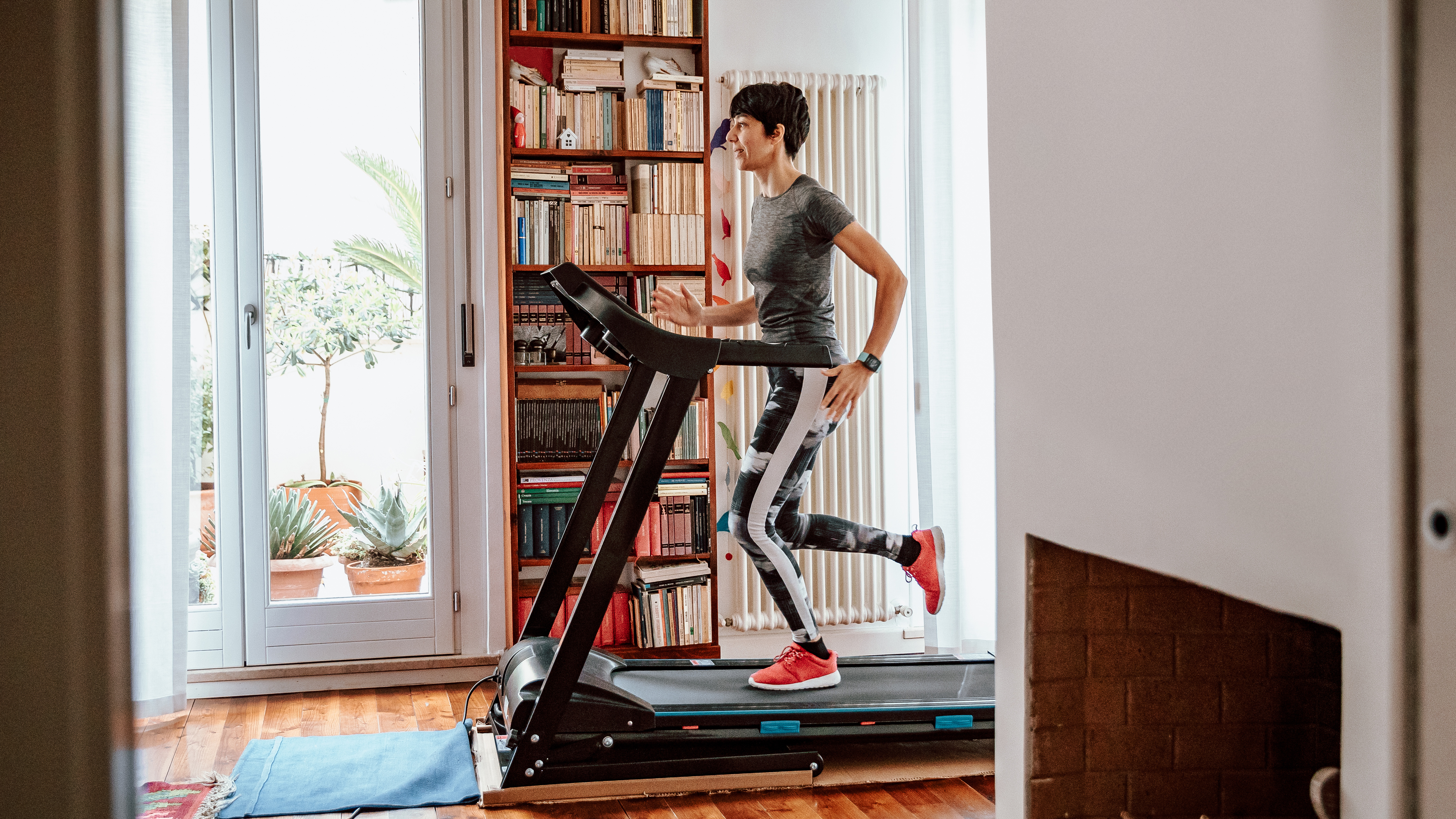 A woman runs on a treadmill in a living room. She is wearing a t-shirt, leggings and sneakers. Behind her we see a book case and a radiator.