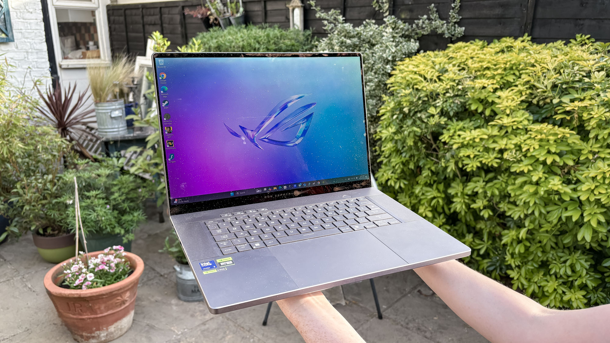 Asus ROG Zephyrus G16 being held with two hands with bushes in background