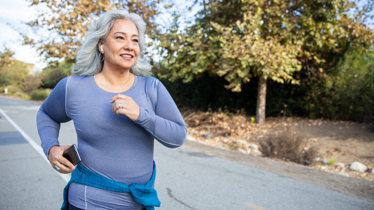 A smiling woman in a long-sleeved top runs on a road. She has a phone in her right hand and a jumper tied around her waist. 