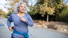 A smiling woman in a long-sleeved top runs on a road. She has a phone in her right hand and a jumper tied around her waist. 