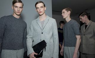 Four male models wearing looks from Jil Sander's collection. They are wearing light and dark grey trousers, tops and jackets. One model is holding a bag