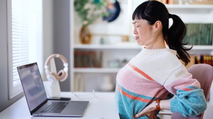 woman wearing a striped jumper sitting at a desk in front of a laptop, holding her lower back and grimacing, sideways onto the camera