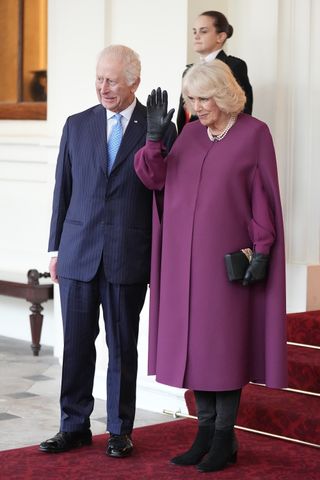 King Charles standing next to Queen Camilla, wearing a purple cape and waving on a red carpeted staircase