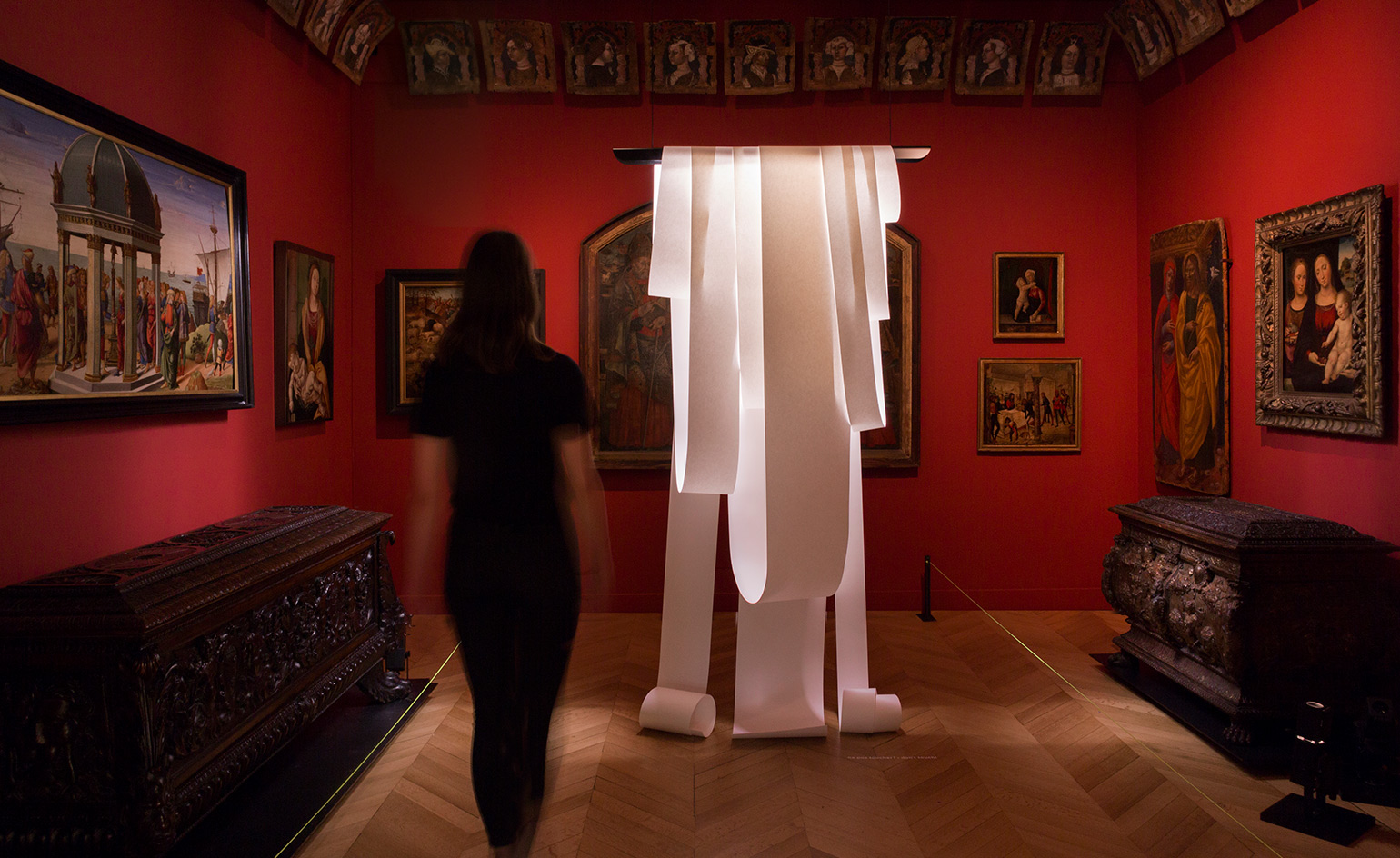 A room with walls painted red, and paintings hung on the walls in different sizes. Antique-looking furniture in deep brown, with elaborate carvings, is set on each side. An art piece that looks like white ribbons with the light coming from the inside of it, is hung from the ceiling.