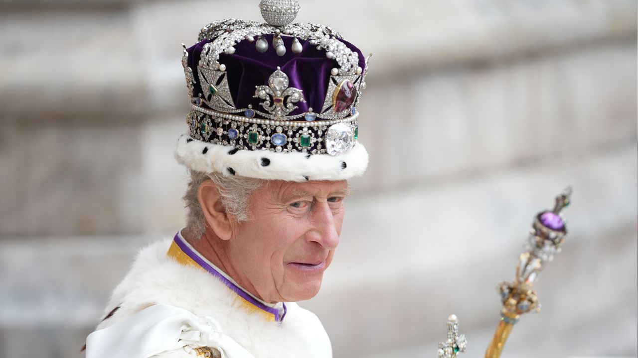 A headshot of King Charles wearing his coronation crown and robes in front of a marble column