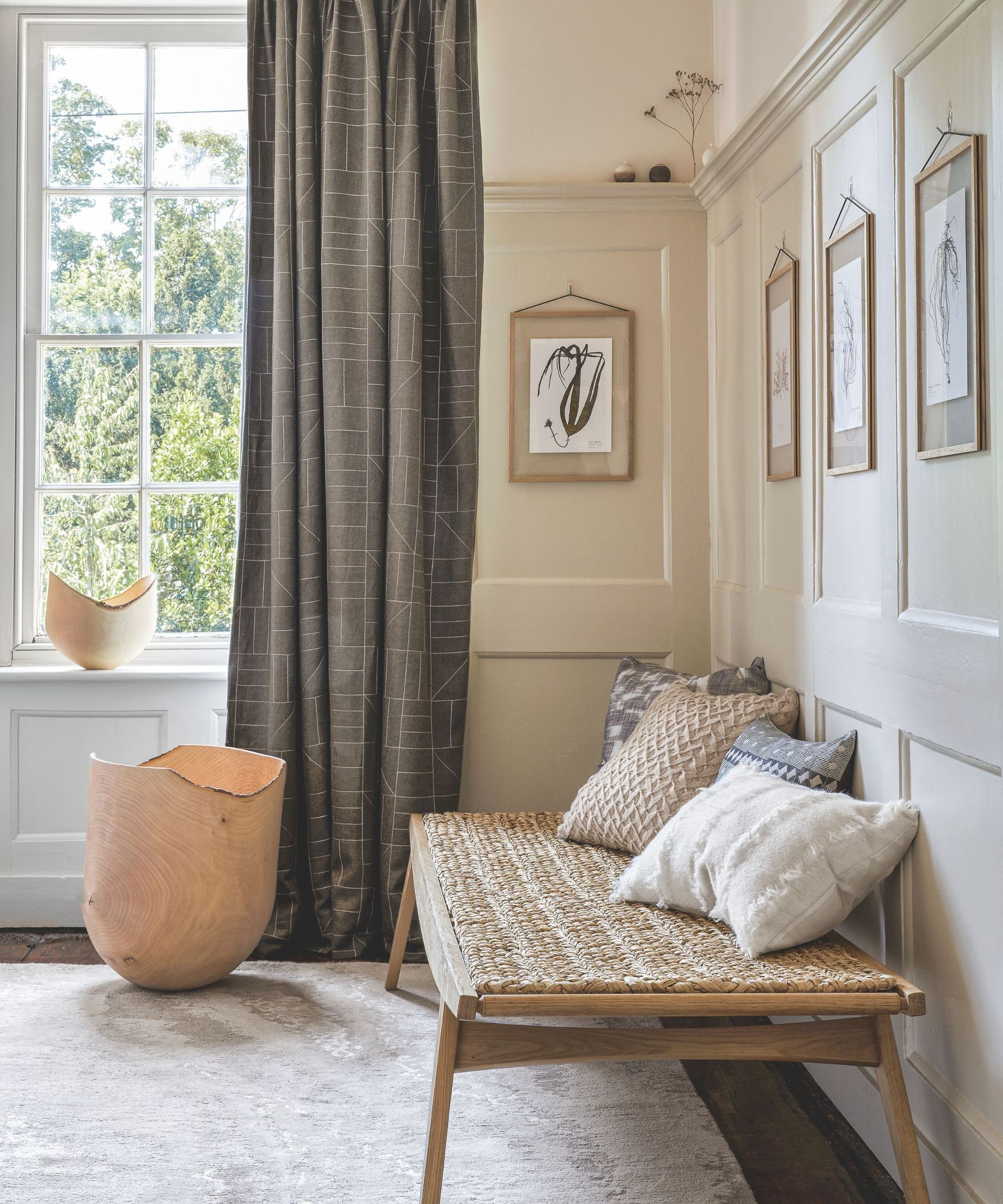 Neutral hallway with large sash window, wood panelled walls painted light beige, a wooden bench with textured cushions below botanical prints hung on the walls, and some statement woodent containers by the window