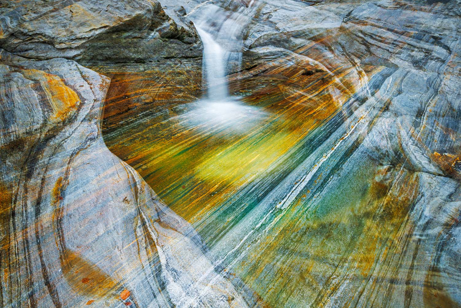 Small waterfall cascading into a colorful, smooth rock pool with streaks of orange, yellow, green, and blue
