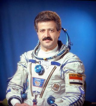 a man with a mustache wearing a white spacesuit poses in front of a blue background