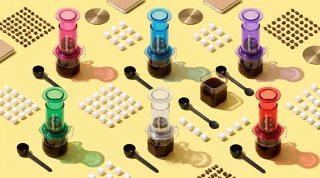 Collection of different colored AeroPress coffee makers lined up in a geometric formation with coffee beans, scoops, and sugar cubes