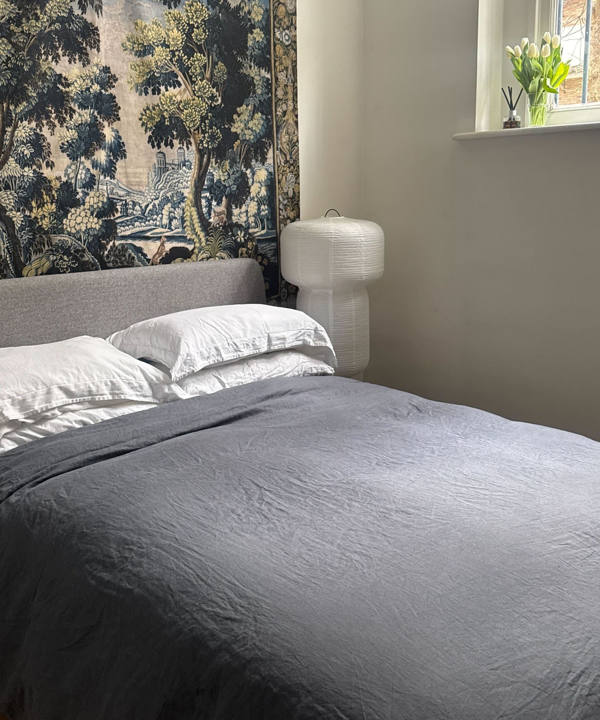 Pottery Barn linen bedding in bedroom with navy bedding, white pillowcases, paper lantern, tapestry, and vase of tulips on windowsill