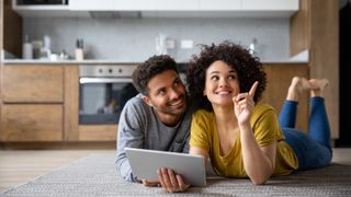 man and woman lying on floor of kitchen with ipad like device in hand looking to right and pointing