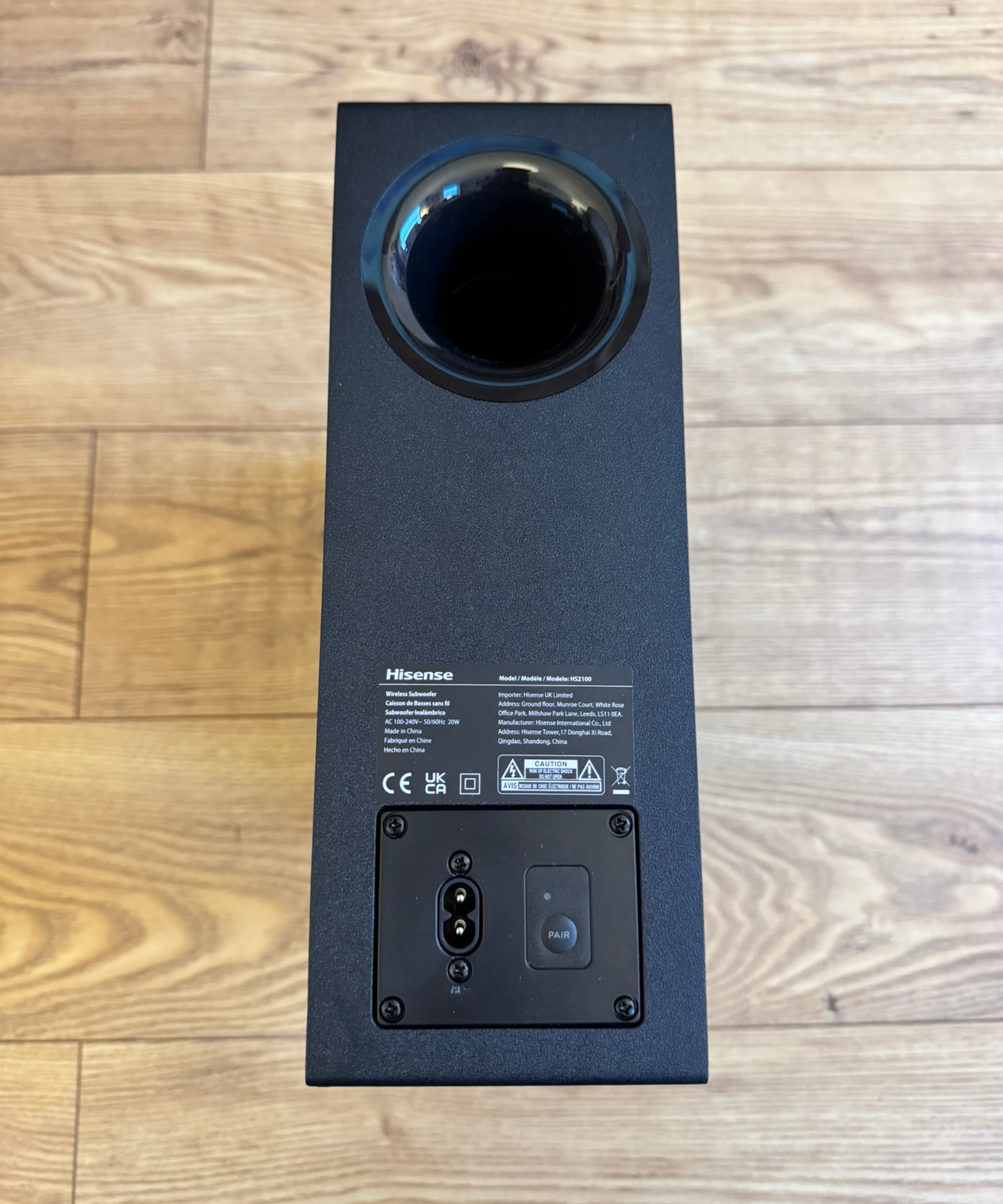Hisense HS2100 subwoofer laid vertically on wooden floor, showing power cord input, 'pair' button, and Hisense manufacture details.
