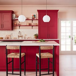 red and pink kitchen with painted ceiling, red cabinets, white worktops, wicker and black painted wood bar stools, brass antique tap, open shelving, white globe pendants