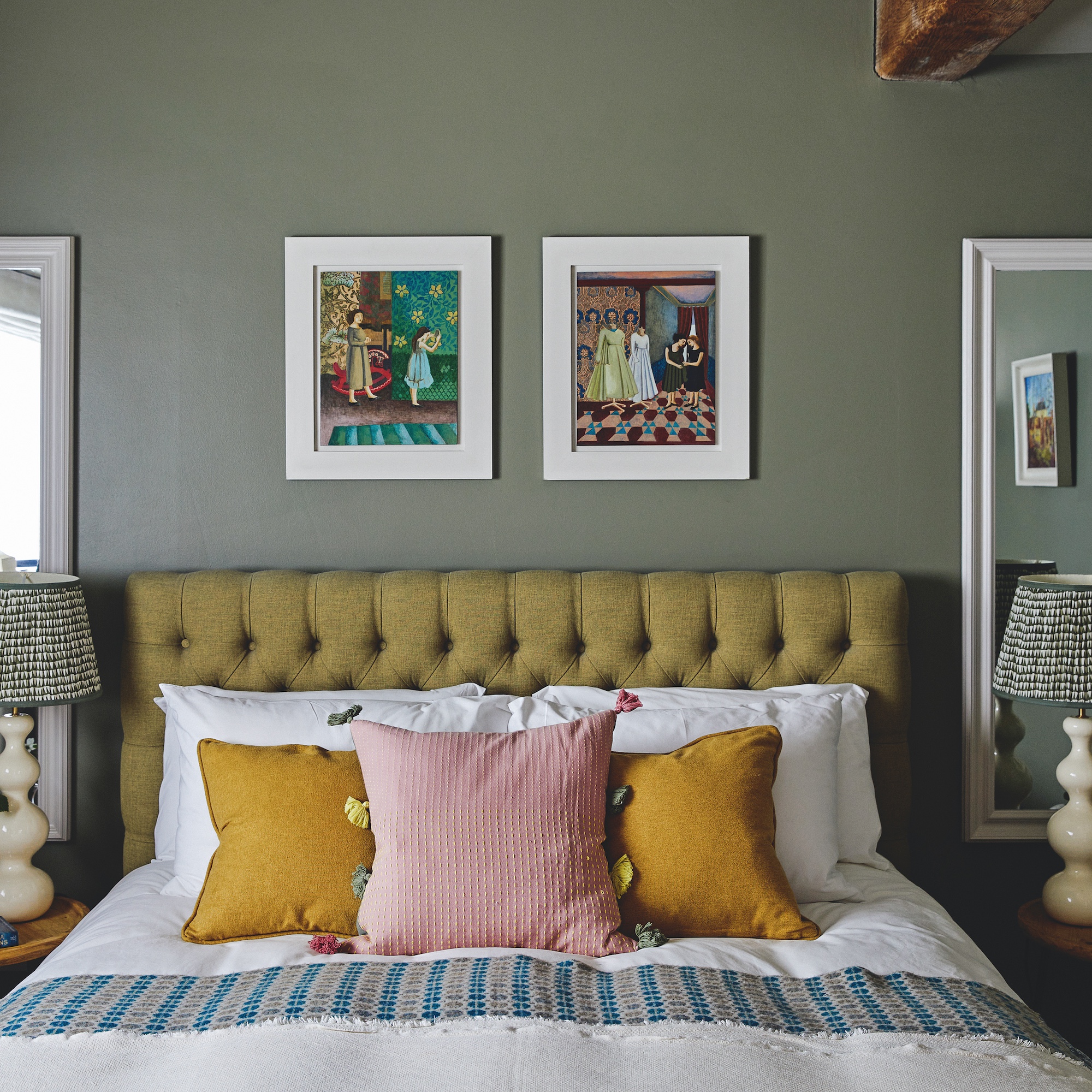 detail of bedrom with sage green walls and upholstered bed in olive green, cushions on bed in pink and ochre, with artworks on wall behind bed