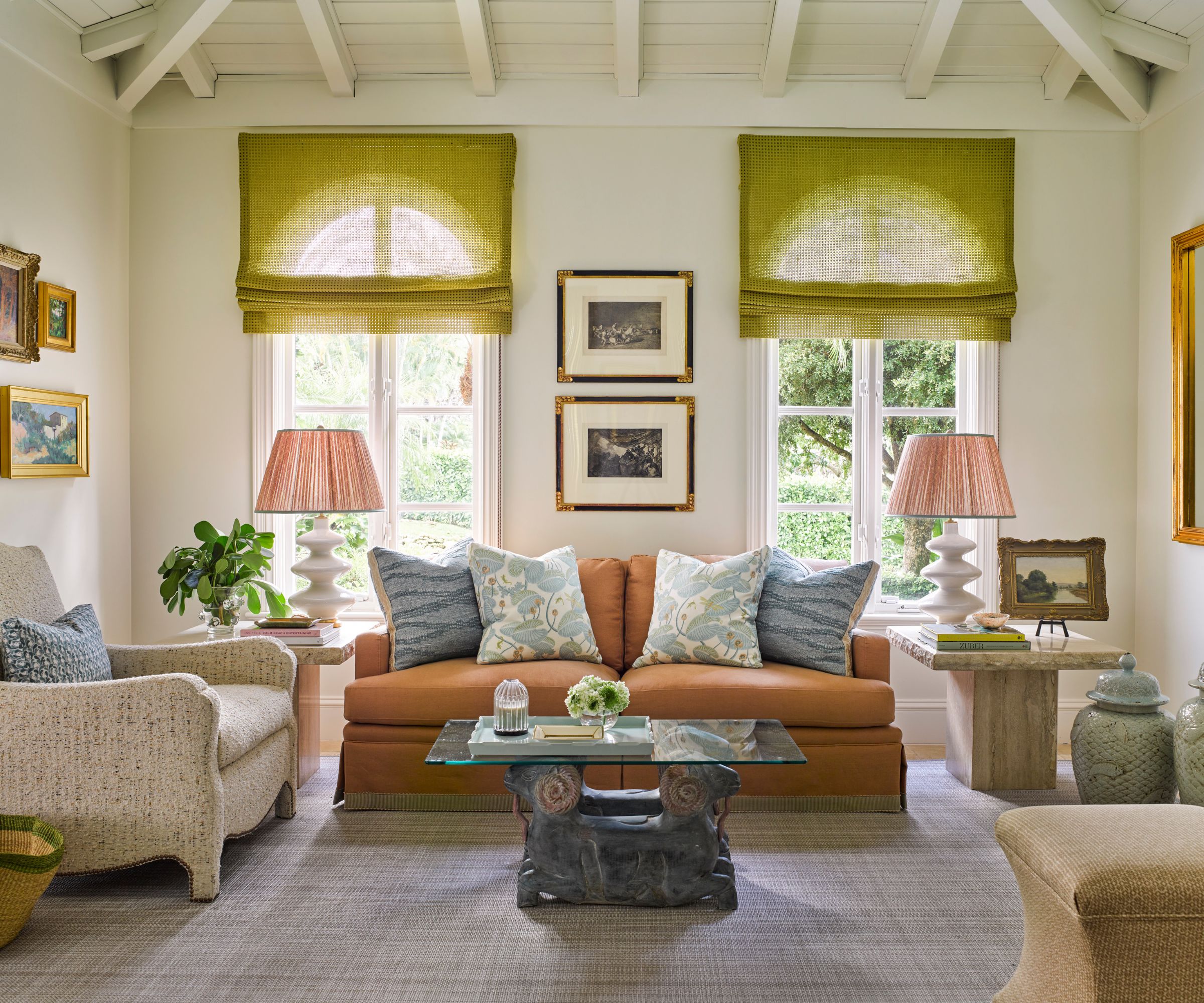 A large white living room with two tall arched windows with lime green sheer roman blinds, a terracotta-colored sofa with pale blue cushions, and two matching pink lampshades either side of the sofa.