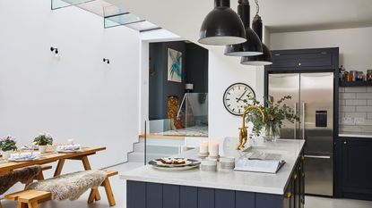 Where should a refrigerator be placed? Kitchen and dining area with concrete floor, black Shaker style kitchen island and units, white metro tiles and black pendant lamps, wooden dining table and benches under glass ceiling.