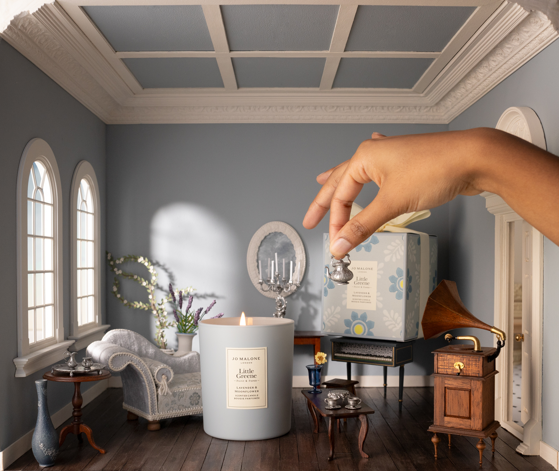 Image of a miniature blue living room with miniature antique furniture. There is a hand coming in from the side holding a mini silver teapot. There is a regular-sized candle in the middle of the room.