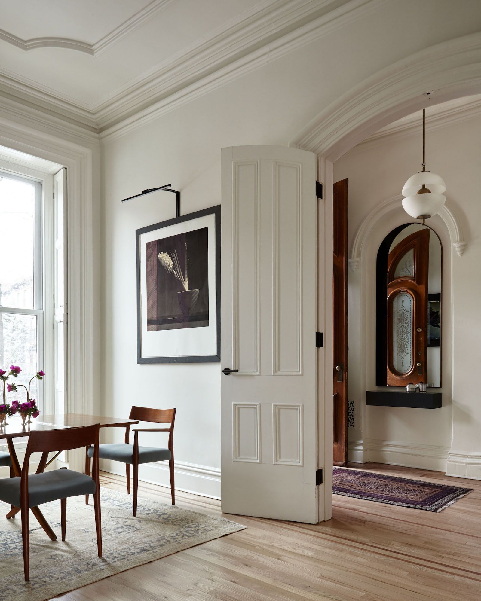 A grand white-walled dining space with an arched doorway leading to an entrance hall with an arched mirror showing an open wooden door
