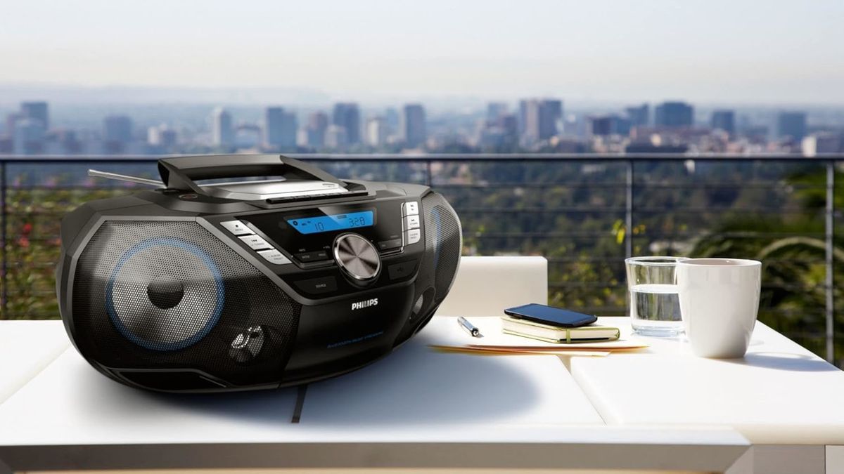 The PHILIPS AZB798T boombox on a table outdoors, beside a coffee cup and glass. In the background there&#039;s a view of a city.